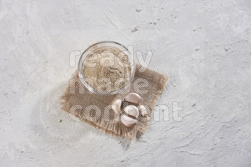 A glass bowl full of garlic powder placed on burlap fabric with garlic cloves on a textured white flooring