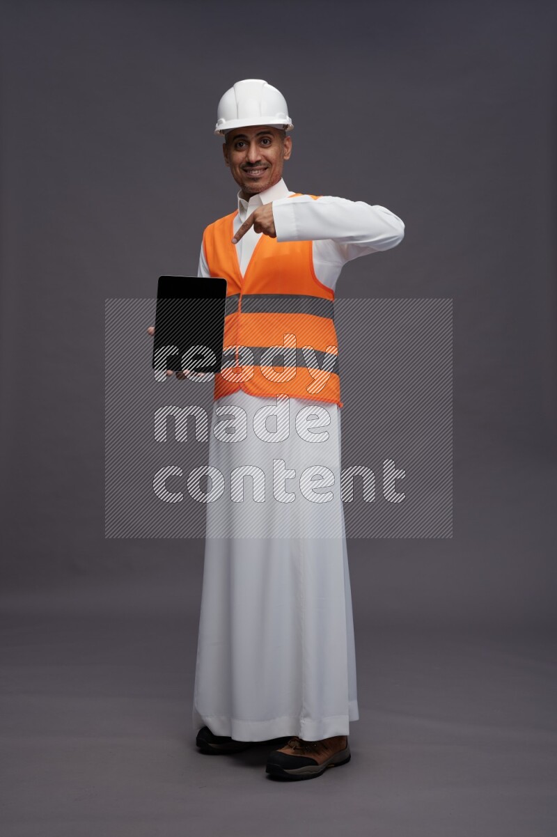 Saudi man wearing thob with engineer vest standing showing tablet to camera on gray background