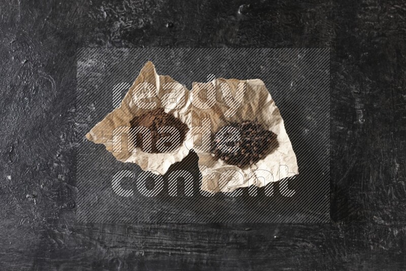 Cloves powder and cloves in 2 crumpled pieces of paper on a textured black flooring