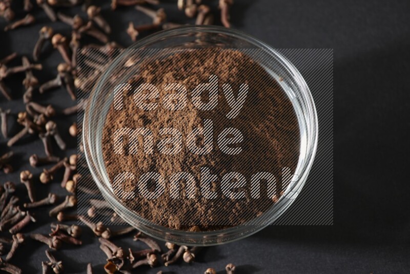 وعاء زجاجي ممتلئ ببودرة القرنفل مع حبوب القرنفل المتناثرة على أرضية سوداء