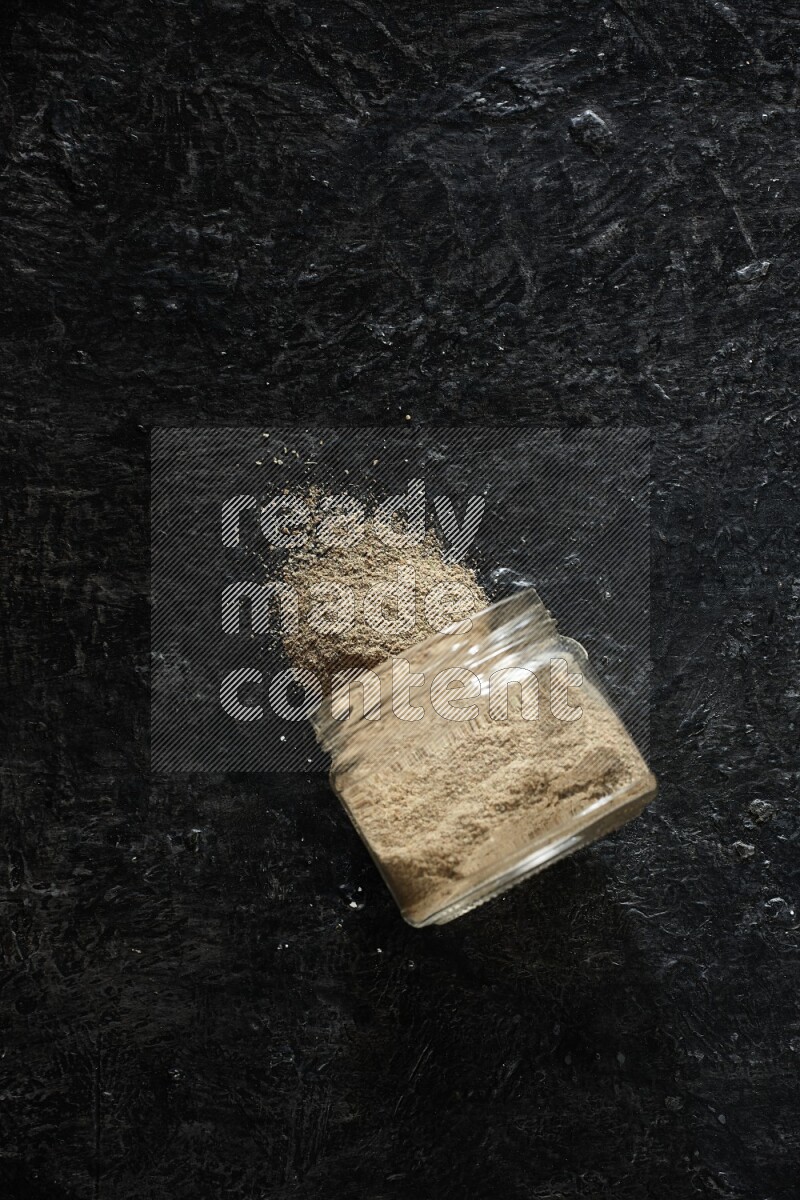 A flipped glass jar full of cardamom powder and powder spilled out of it on textured black flooring