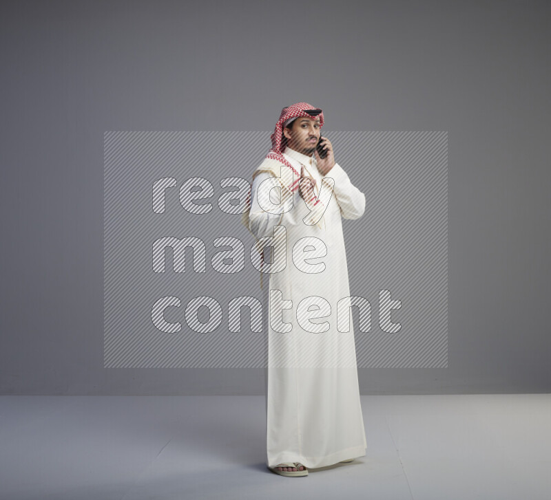 A Saudi man standing wearing thob and red shomag talking on phone on gray background
