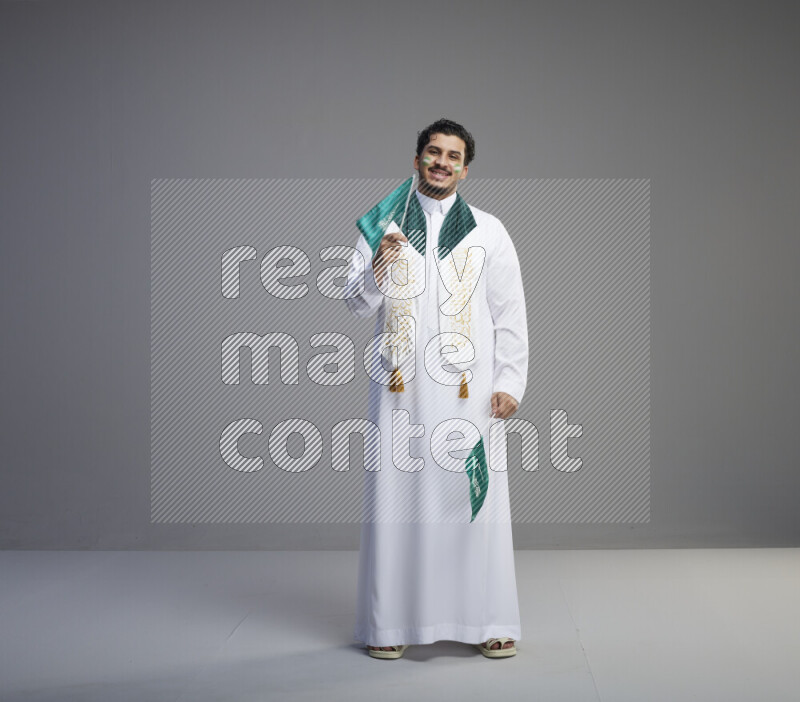 A Saudi man standing wearing thob and saudi flag scarf with face painting holding small Saudi flag on gray background