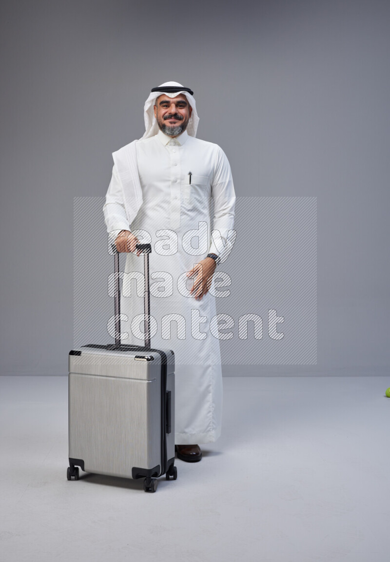 Saudi man wearing Thob and white Shomag standing holding Travel bag on Gray background