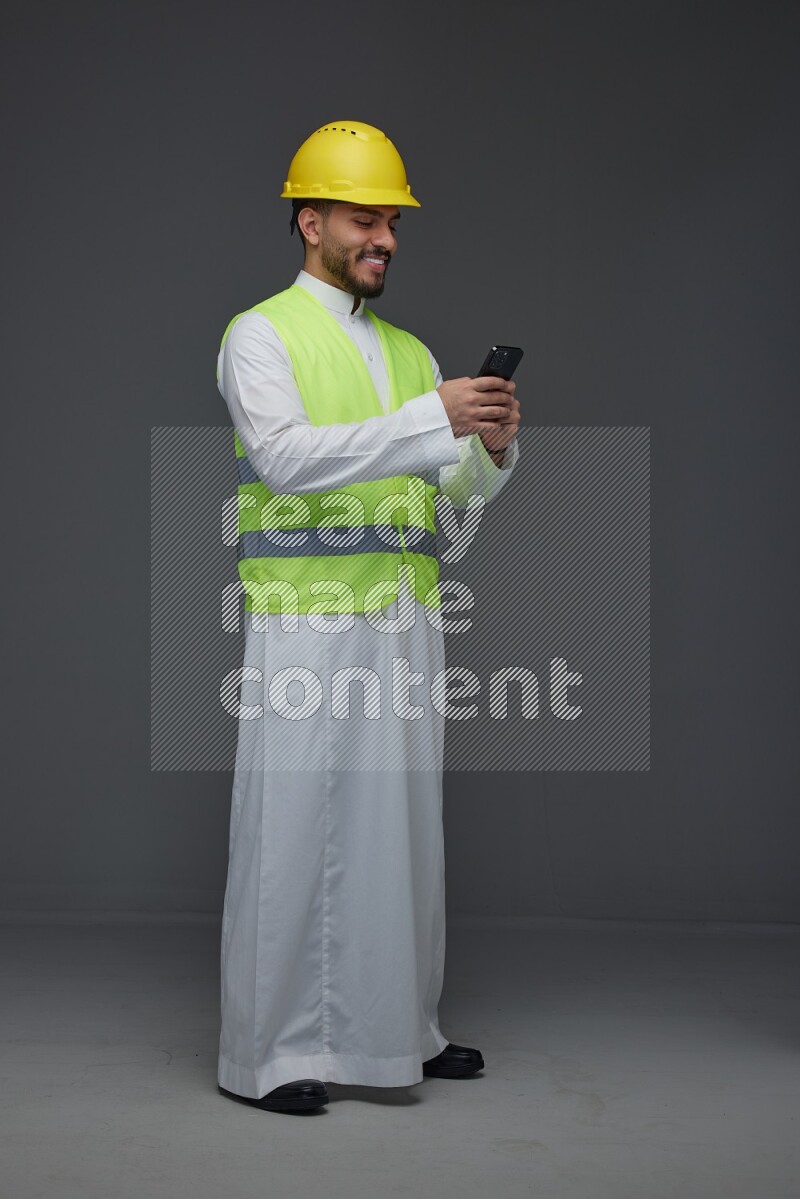 A Saudi man wearing Thobe with a yellow safety vest and white helmet standing and using his phone eye level on a gray background