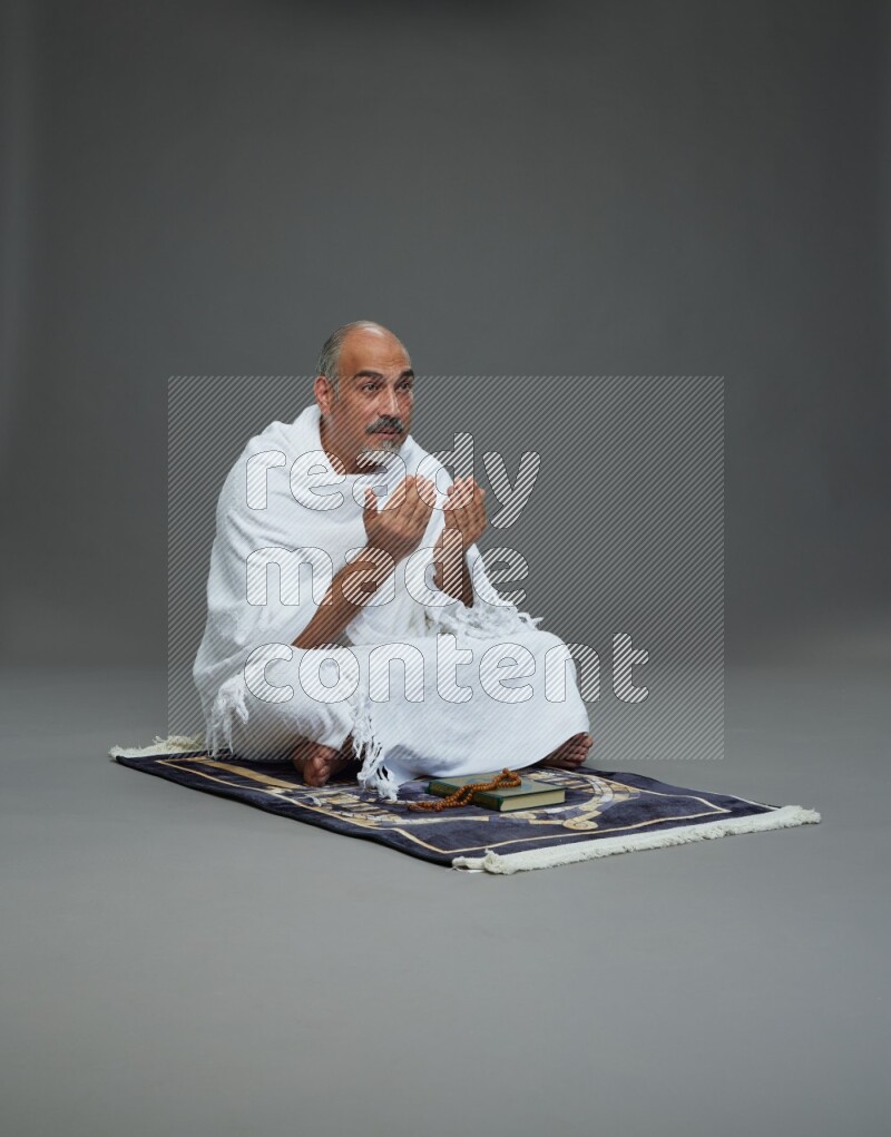 A man wearing Ehram sitting on prayer mat dua'a on gray background