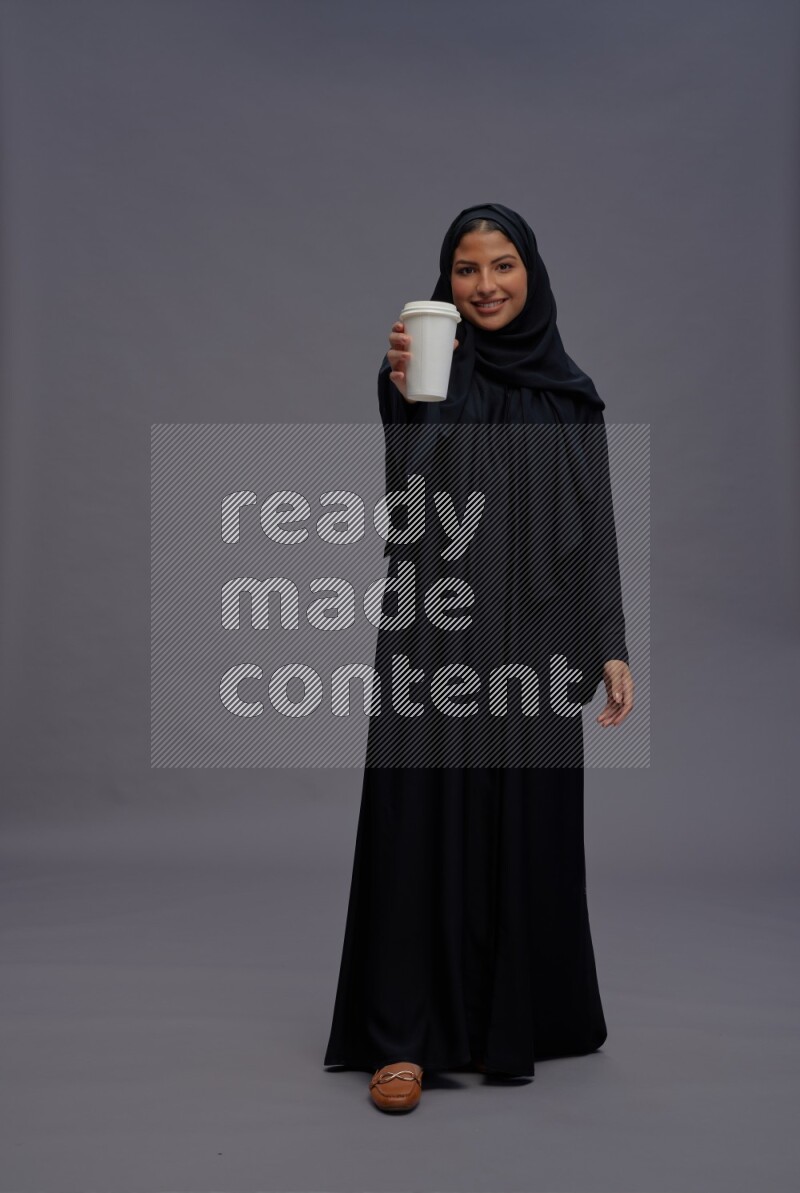 Saudi woman wearing Abaya standing holding paper cup on gray background