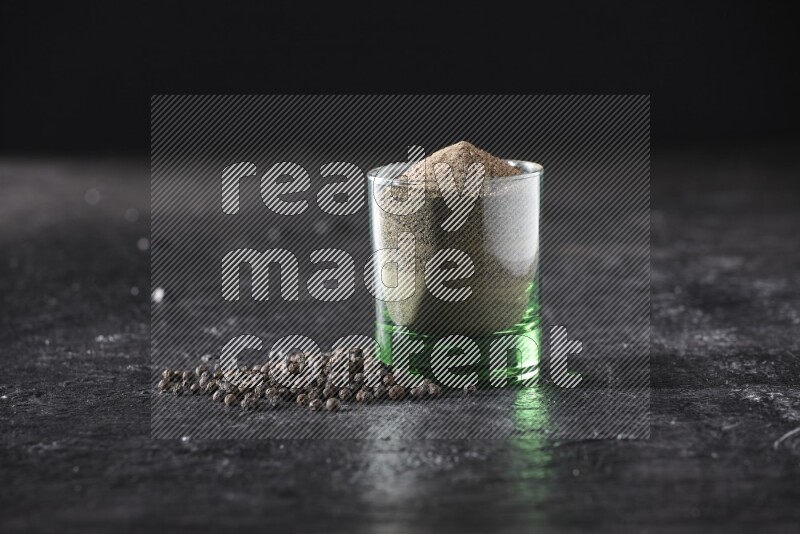 A glass cup full of black pepper powder with beads beside it on a textured black flooring