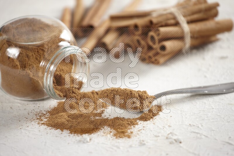 Flipped herbs glass jar full of cinnamon powder with a metal spoon full of powder and cinnamon sticks on a textured white background