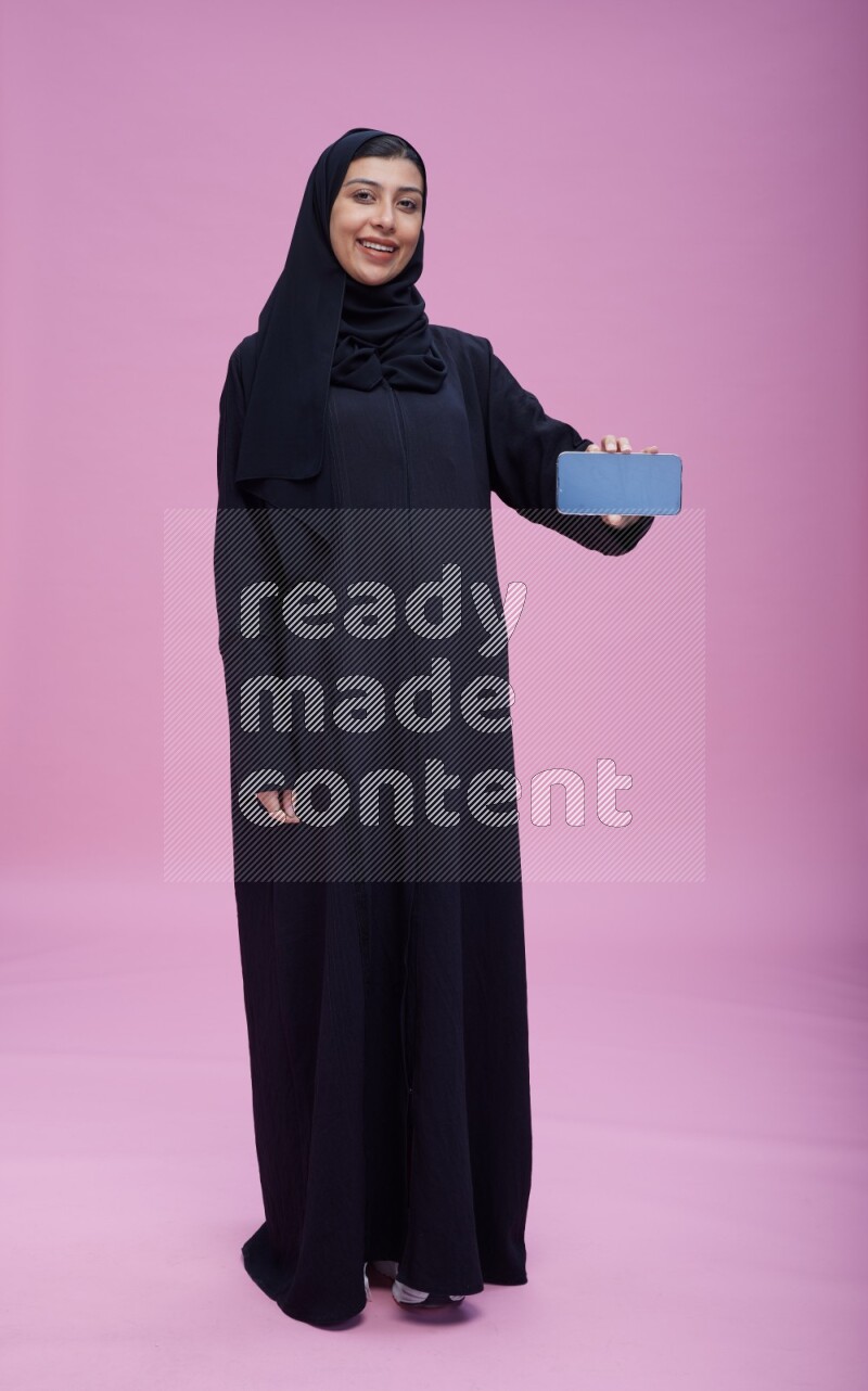 Saudi woman wearing Abaya standing showing phone to camera on pink background
