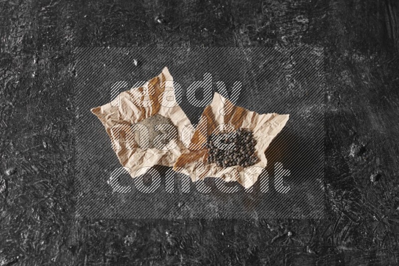 Black pepper and black pepper powder on 2 crumpled paper on a textured black flooring