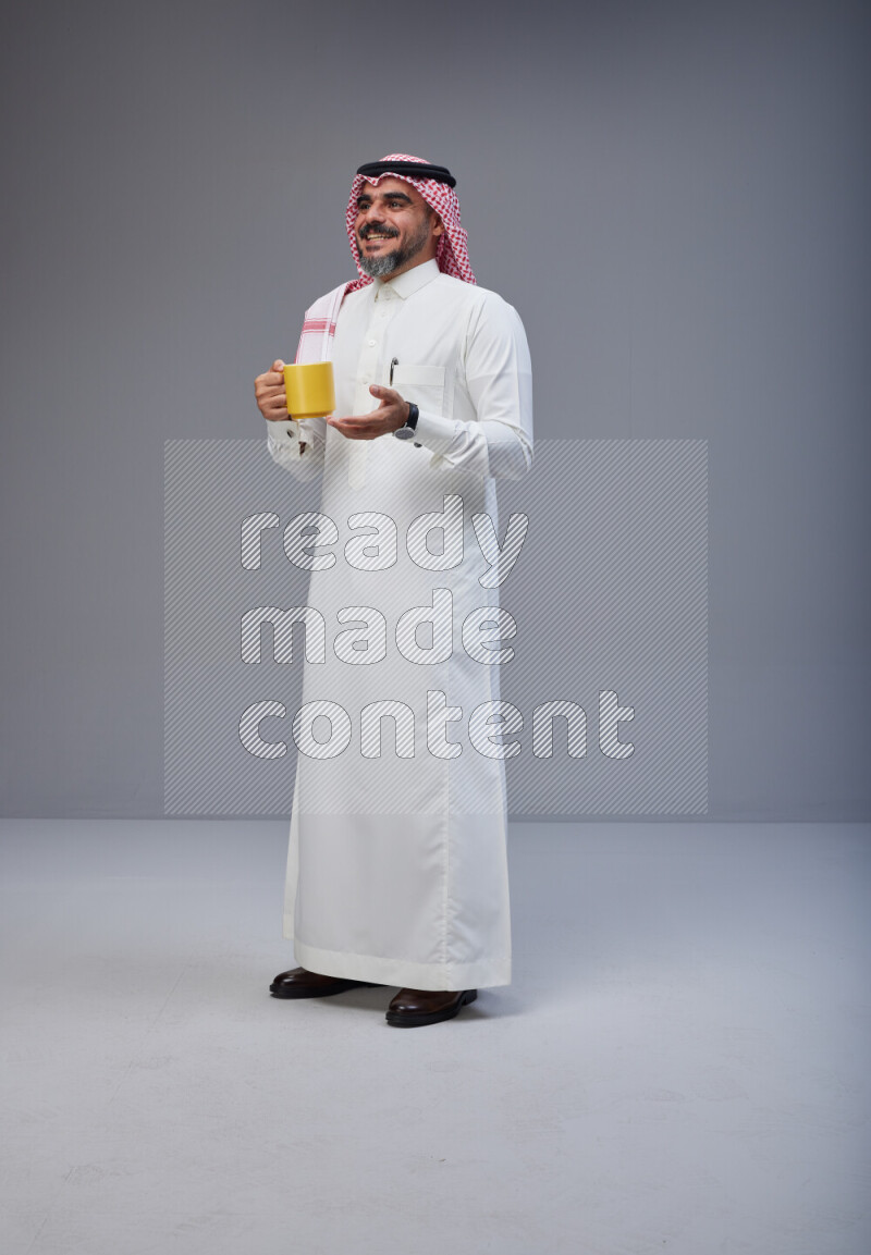 Saudi man Wearing Thob and red Shomag standing holding mug on Gray background