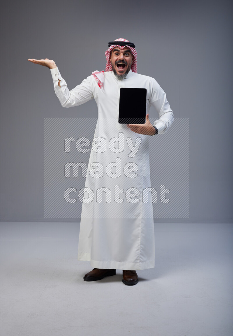 Saudi man Wearing Thob and red Shomag standing showing tablet to camera on Gray background