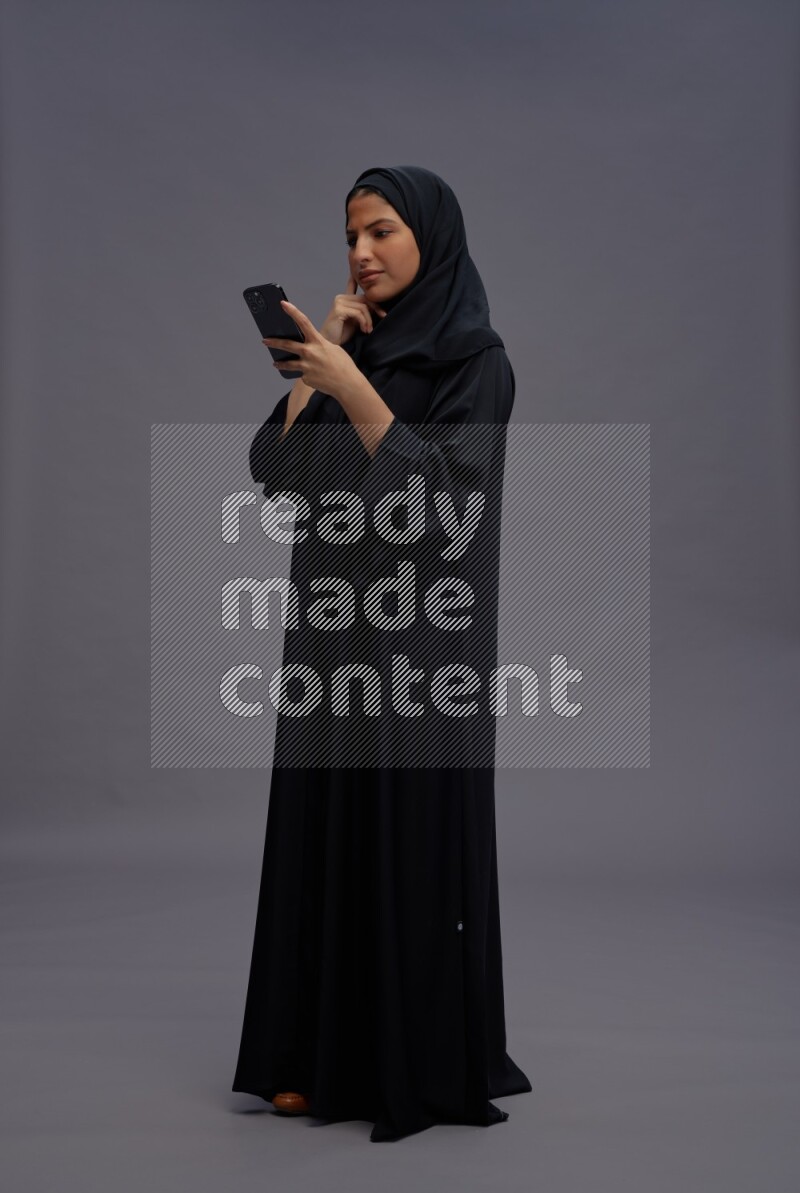 Saudi woman wearing Abaya standing texting on phone on gray background