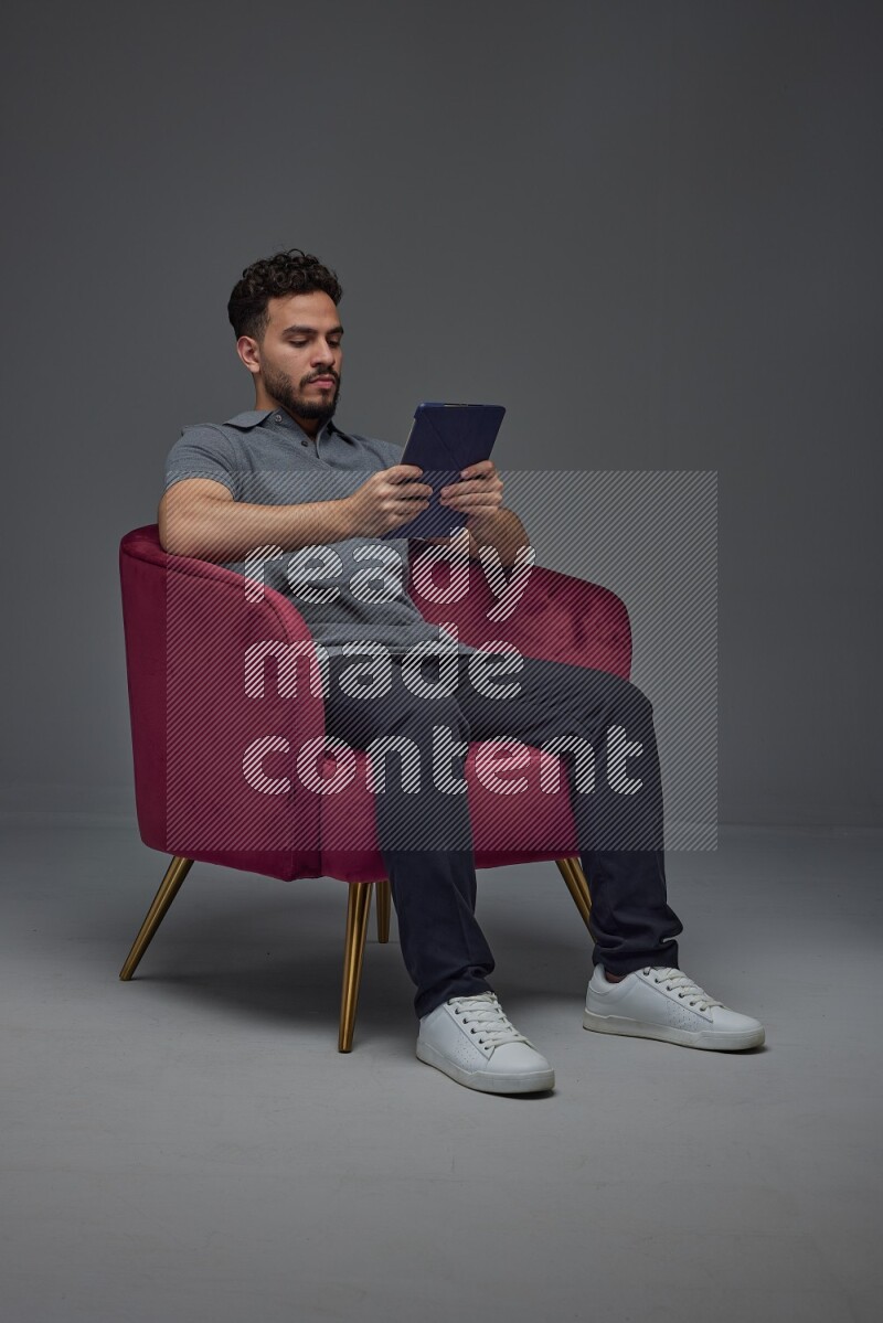 A man wearing casual and using his tablet while sitting on a burgundy chair eye level on a gray background