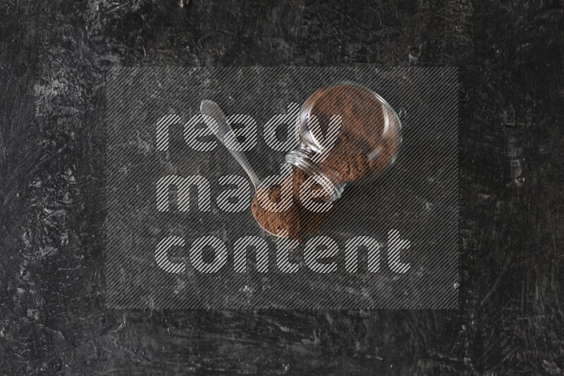 A flipped glass spice jar and a metal spoon full of cloves powder on textured black flooring