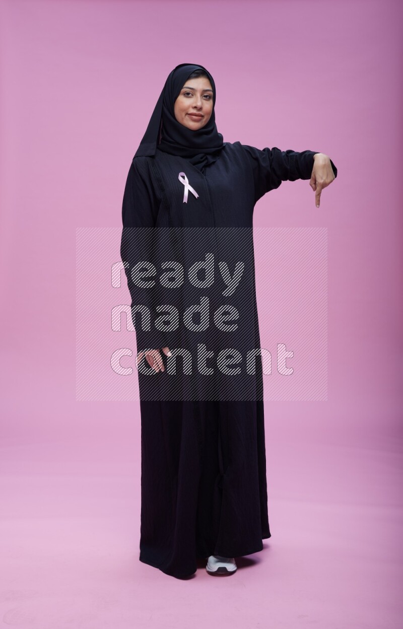 Saudi woman wearing pink ribbon on Abaya standing interacting with the camera on pink background