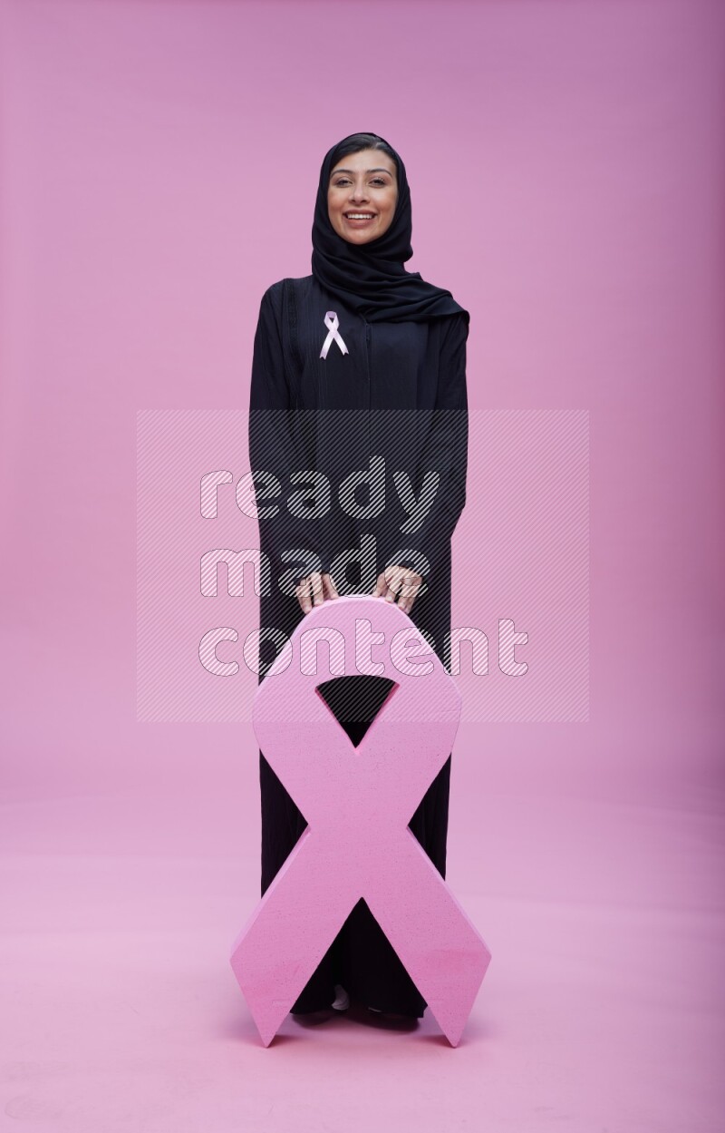 Saudi woman wearing pink ribbon on abaya standing holding awareness ribbon on plain pink background