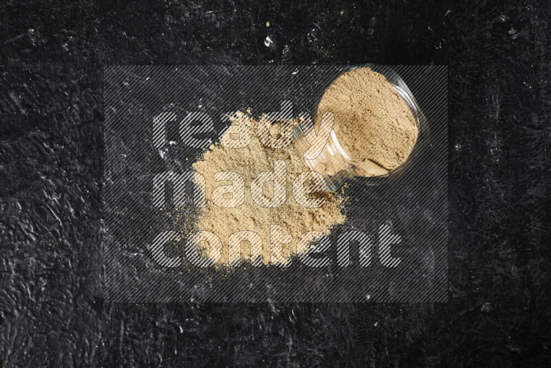 A glass jar full of ground ginger powder flipped with some spilling powder on black background