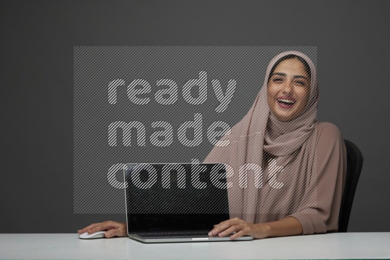 A woman Sitting on her desk  Pointing at her laptop on a Gray Background wearing Brown Abaya with Hijab