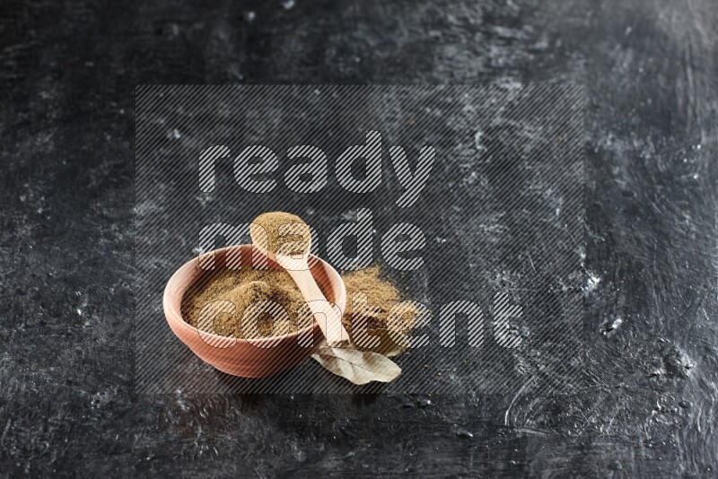 A wooden bowl and spoon full of cumin powder on a textured black flooring