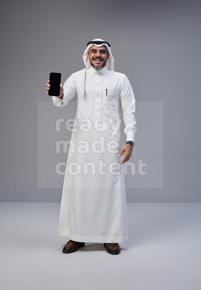 Saudi man Wearing Thob and white Shomag standing showing phone to camera on Gray background