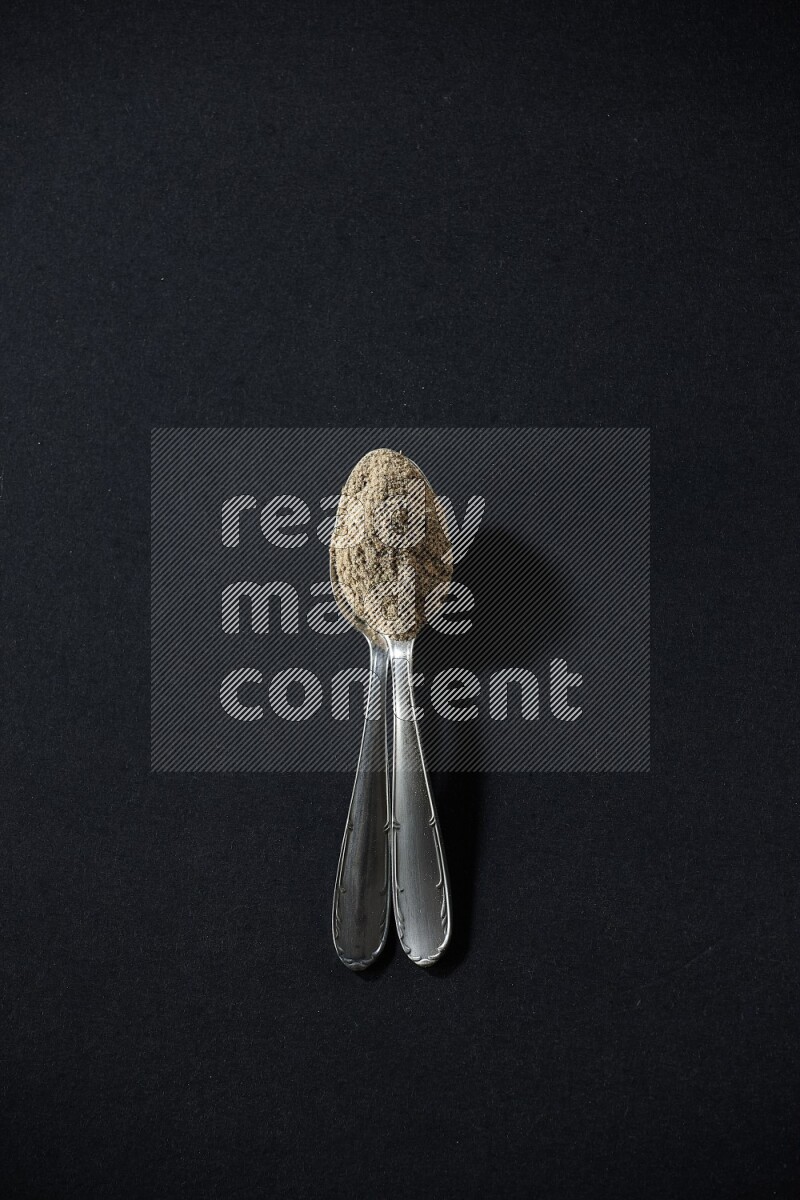 2 Metal spoons full of cardamom powder on black flooring