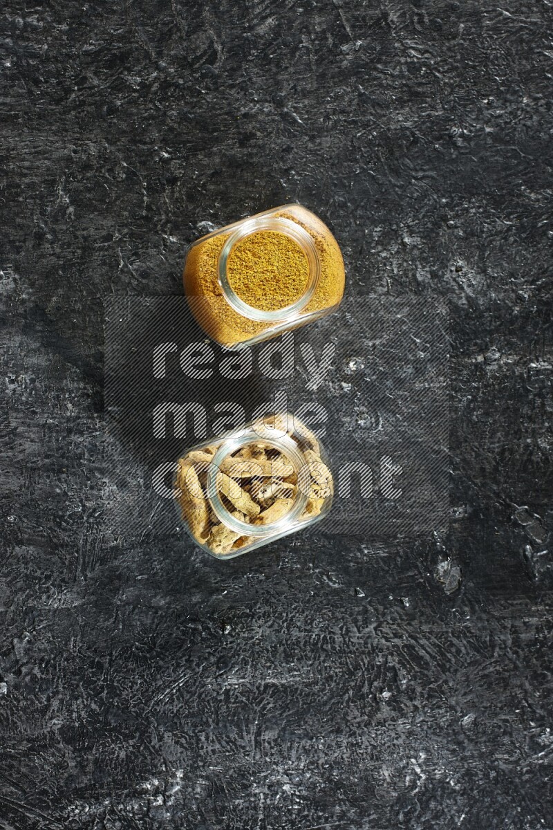 2 glass spice jars full of turmeric powder and dried whole fingers on textured black flooring
