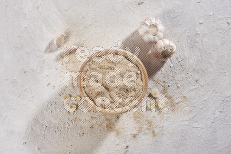 A wooden bowl full of garlic powder and beside it garlic cloves on a textured white flooring in different angles