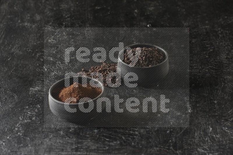 2 Black pottery bowls full of cloves and the other full of cloves powder on textured black flooring