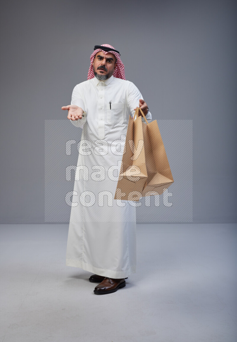 Saudi man Wearing Thob and red Shomag standing holding shopping bag on Gray background