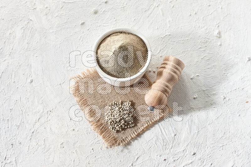 White pottery bowl full of white pepper powder set on a burlap piece of fabric with pepper beads and wooden pepper grinder on textured white flooring