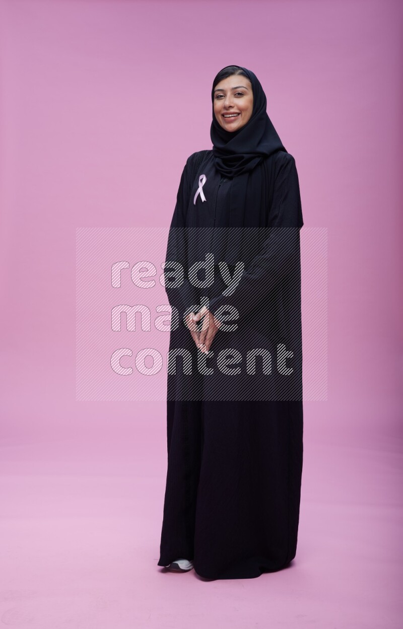 Saudi woman wearing pink ribbon on Abaya standing interacting with the camera on pink background