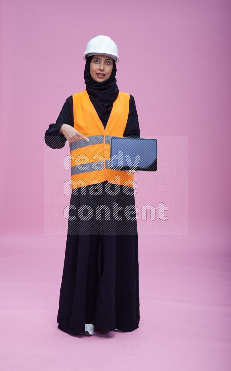 Saudi woman wearing Abaya with engineer vest and helmet standing showing tablet to camera on pink background