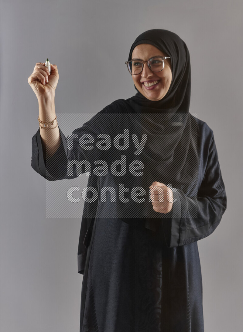 Woman in black abaya, hijab and glasses holding a marker pen to write on a transparent board with different reactions and expressions on grey background