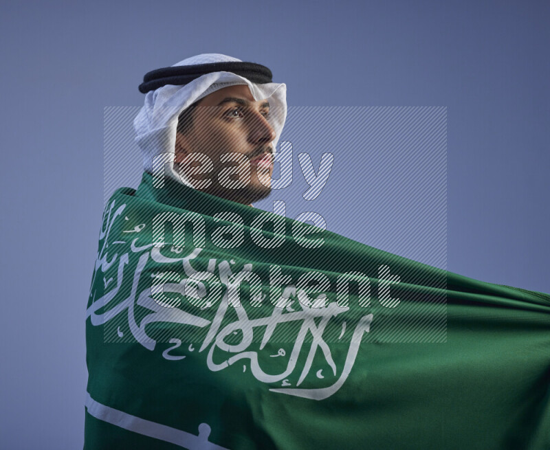 A close-up shot of Saudi man wearing thob and white shomag wrapping big Saudi flag on gray background