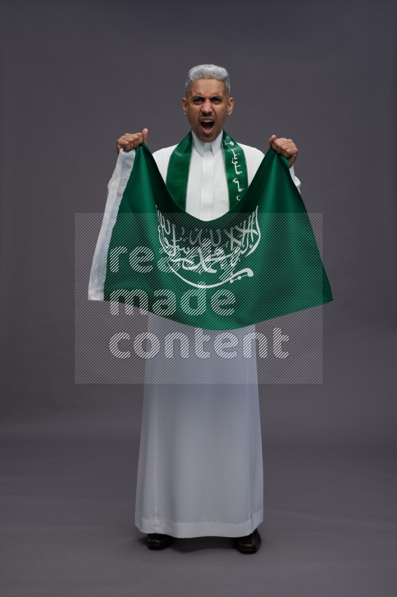 Saudi man wearing thob standing holding Saudi flag on gray background