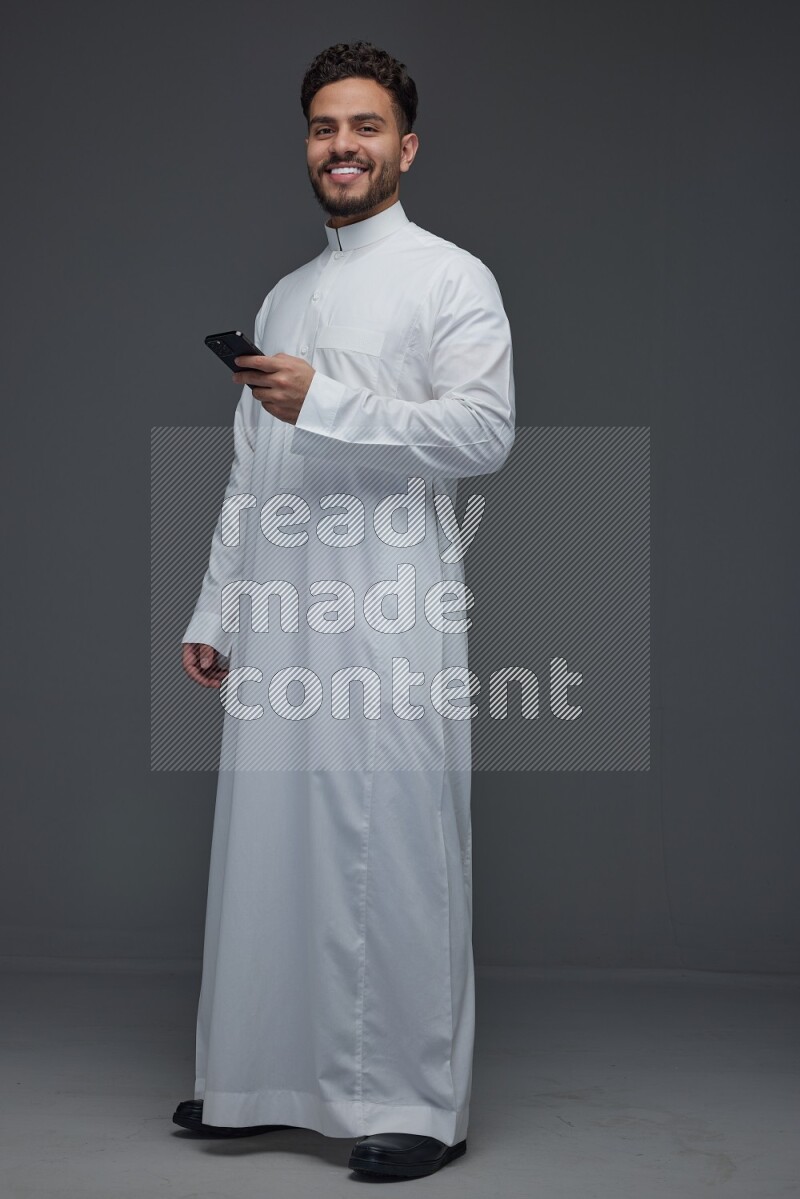 A Saudi man wearing Thobe standing and using his phone eye level on a gray background