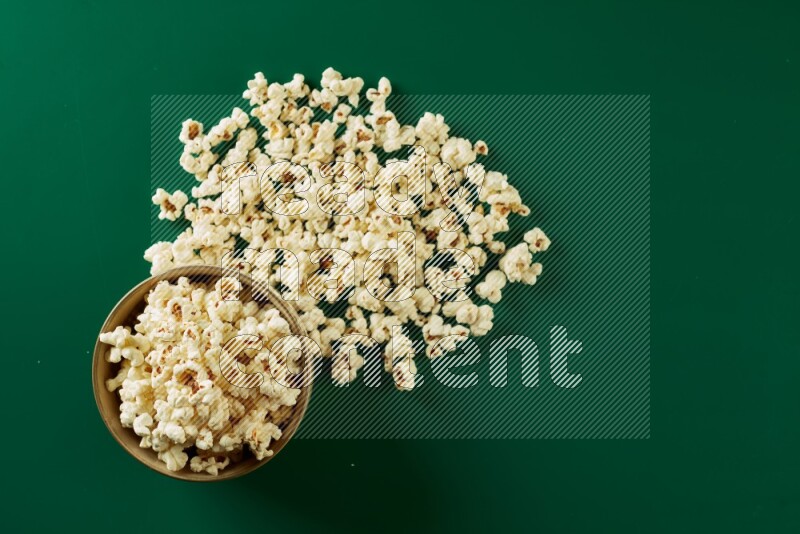 An off white ceramic bowl full of popcorn with popcorn beside it on a green background in a top view shot