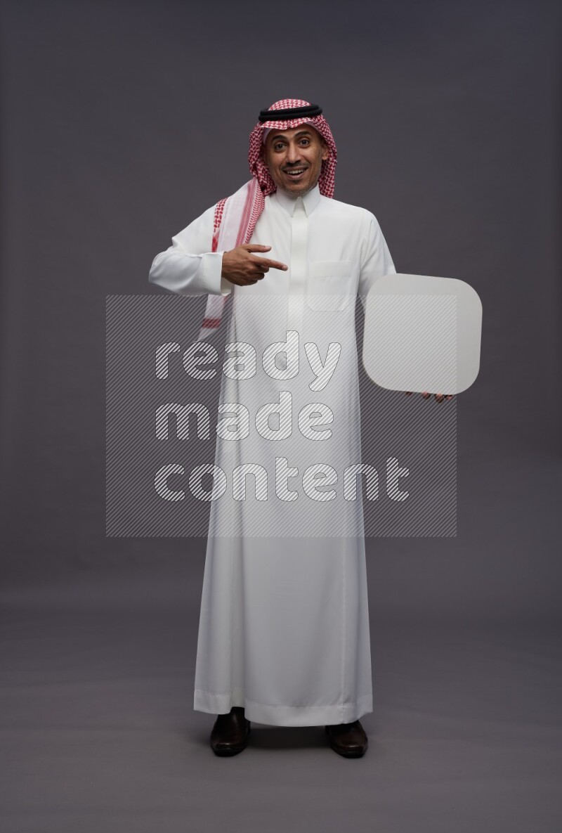 Saudi man wearing thob and shomag standing holding social media sign on gray background