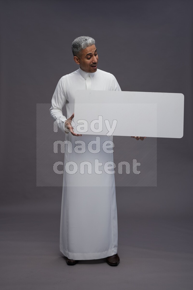 Saudi man wearing thob standing holding board on gray background