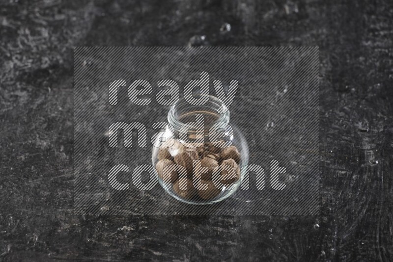 A glass spice jar full of whole nutmeg seeds on a textured black flooring