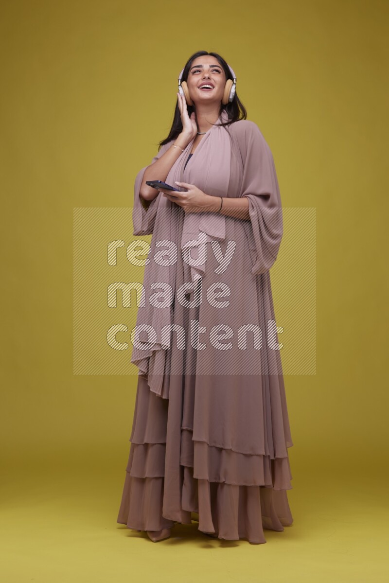A Saudi woman Listing to Music on a Yellow Background wearing Brown Abaya