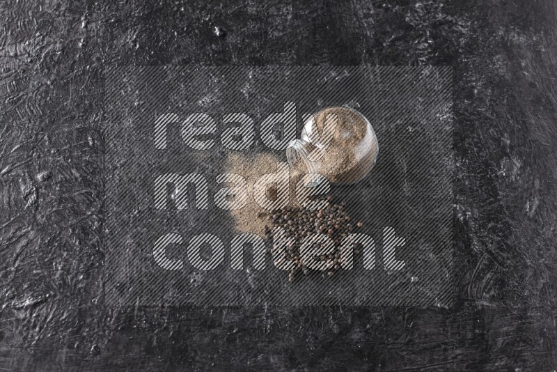 بودرة الفلفل الأسود في وعاء زجاجي مقلوب مع حبوب الفلفل الأسود بجانبه علي خلفية سوداء