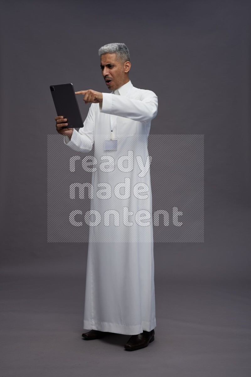 Saudi man wearing thob with neck strap employee badge standing working on tablet on gray background