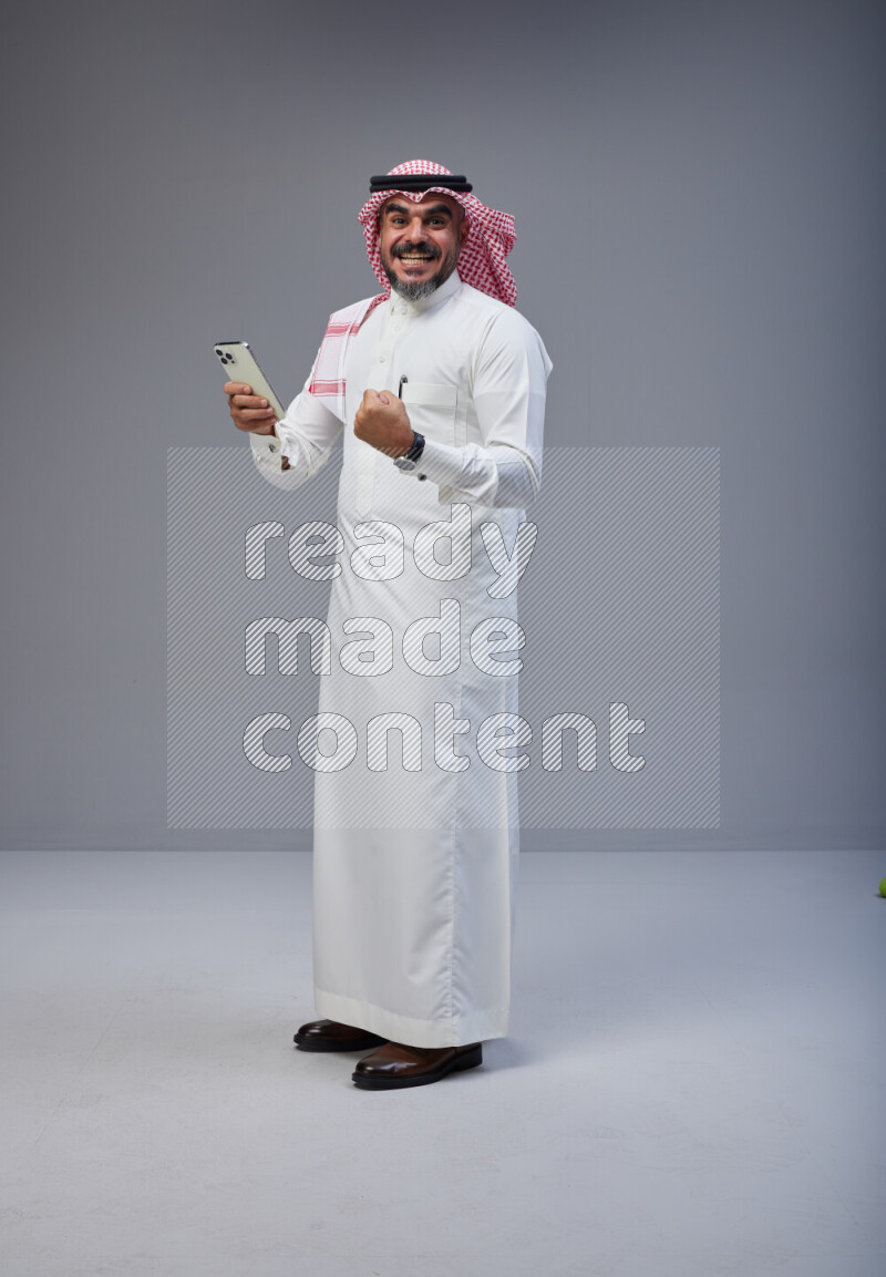 Saudi man Wearing Thob and red Shomag standing texting on phone on Gray background