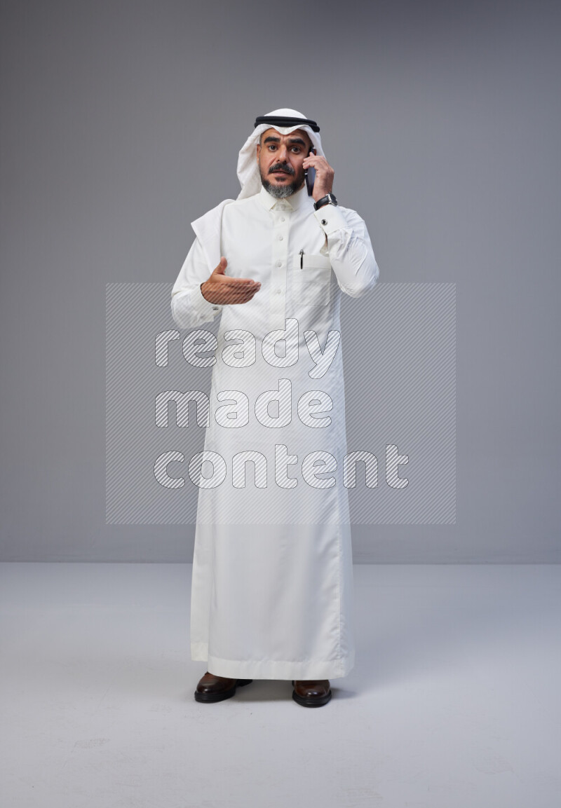 Saudi man Wearing Thob and white Shomag standing talking on phone on Gray background