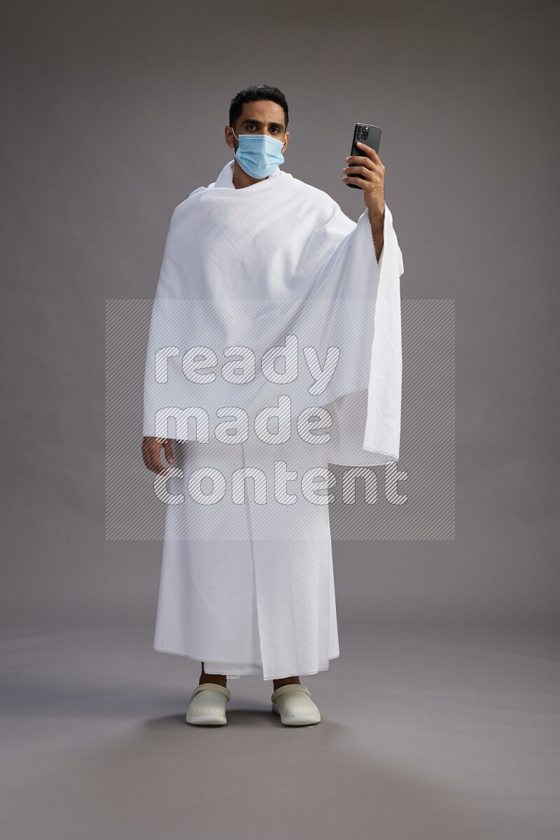 A man wearing Ehram with face mask Standing taking selfie on gray background