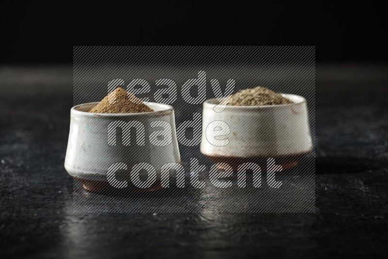 2 beige bowls, one full of cumin powder and the other full of seeds on a textured black flooring