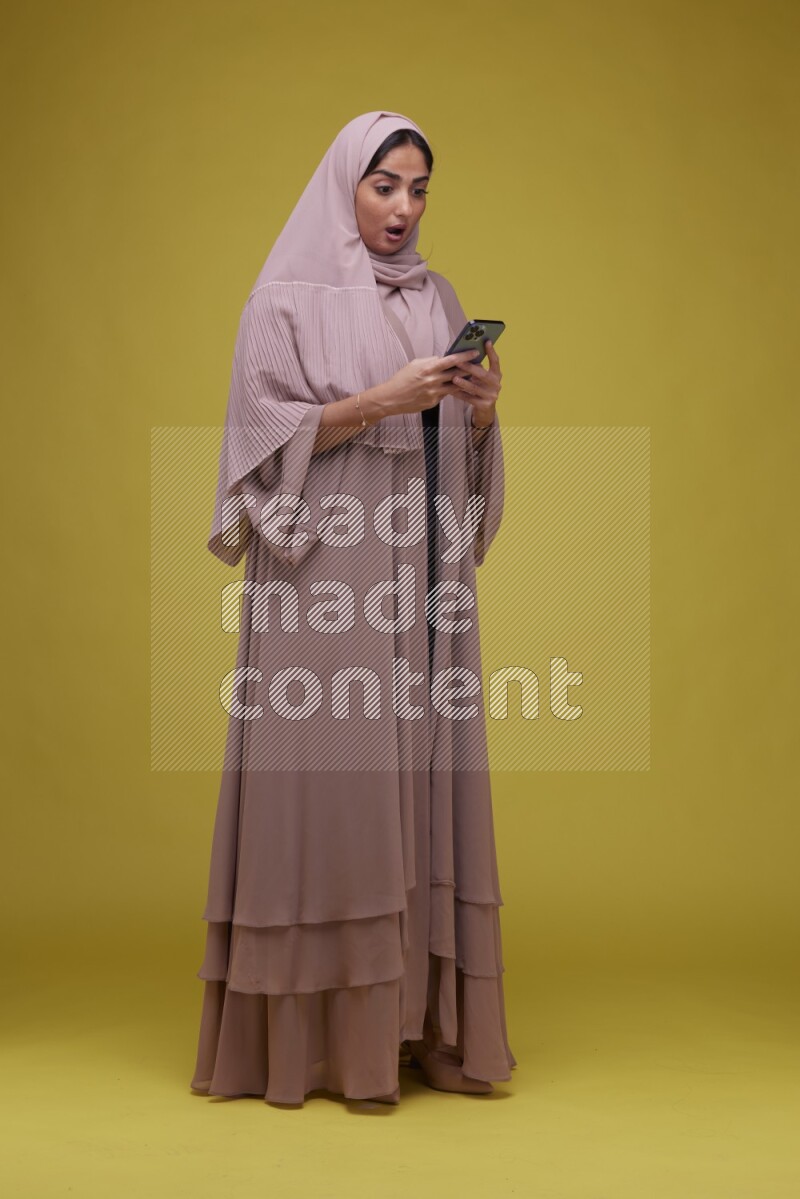 A woman Texting on her phone on a Yellow Background wearing Brown Abaya with Hijab